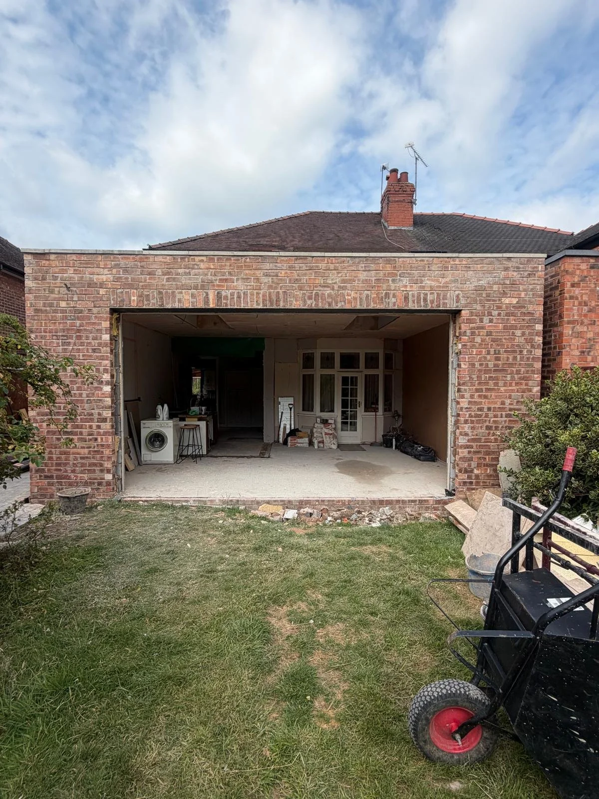 Construction site in backyard showing an unfinished brick wall with large opening, tools, and stacked materials inside, with a grassy area and a wheelbarrow in the foreground.