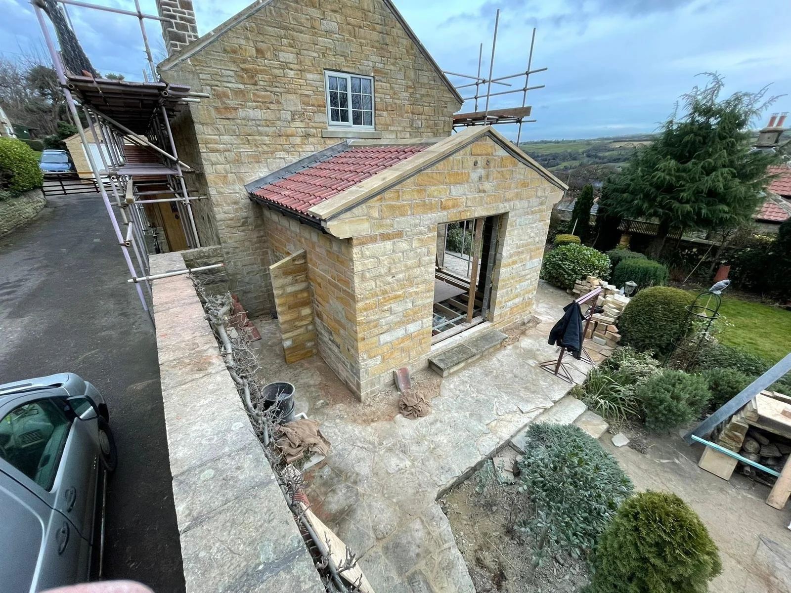 A house under construction with scaffolding around it. The house is built with tan stone bricks and has a small extension with a red tile roof. There is a driveway with a parked gray car on the left, and a garden with various shrubs and trees on the 