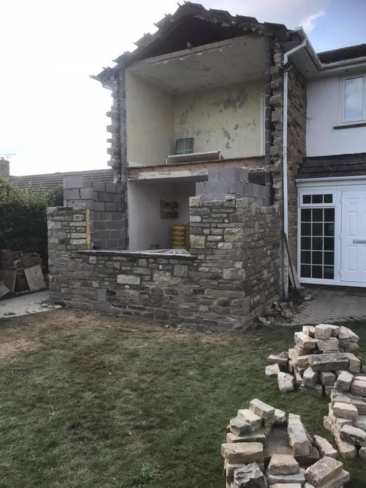 A house undergoing renovation with a partially built stone wall on the ground floor and a missing second floor wall, showing the interior. Piles of bricks are on the grass in the foreground.