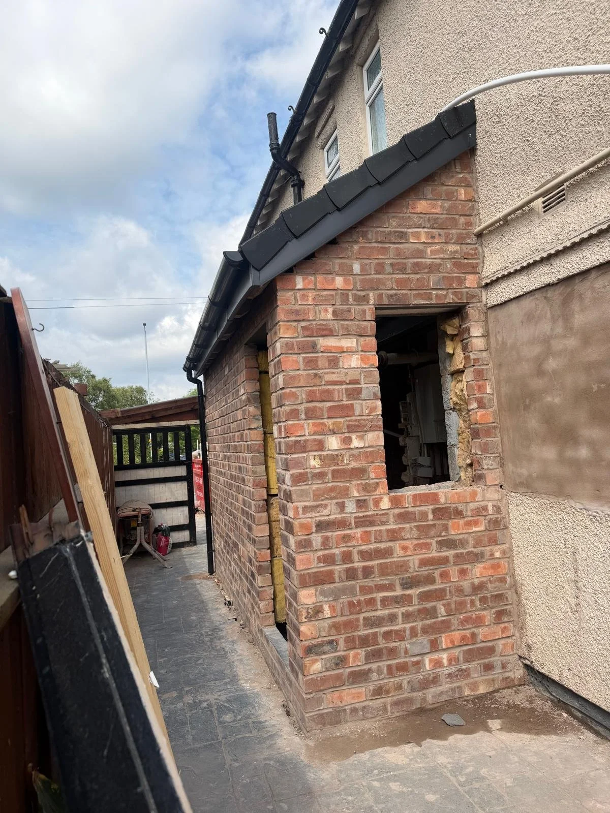 A construction site showing a brick extension being built onto a house, with some construction tools and materials visible. The house has a small window and a partially completed exterior wall.