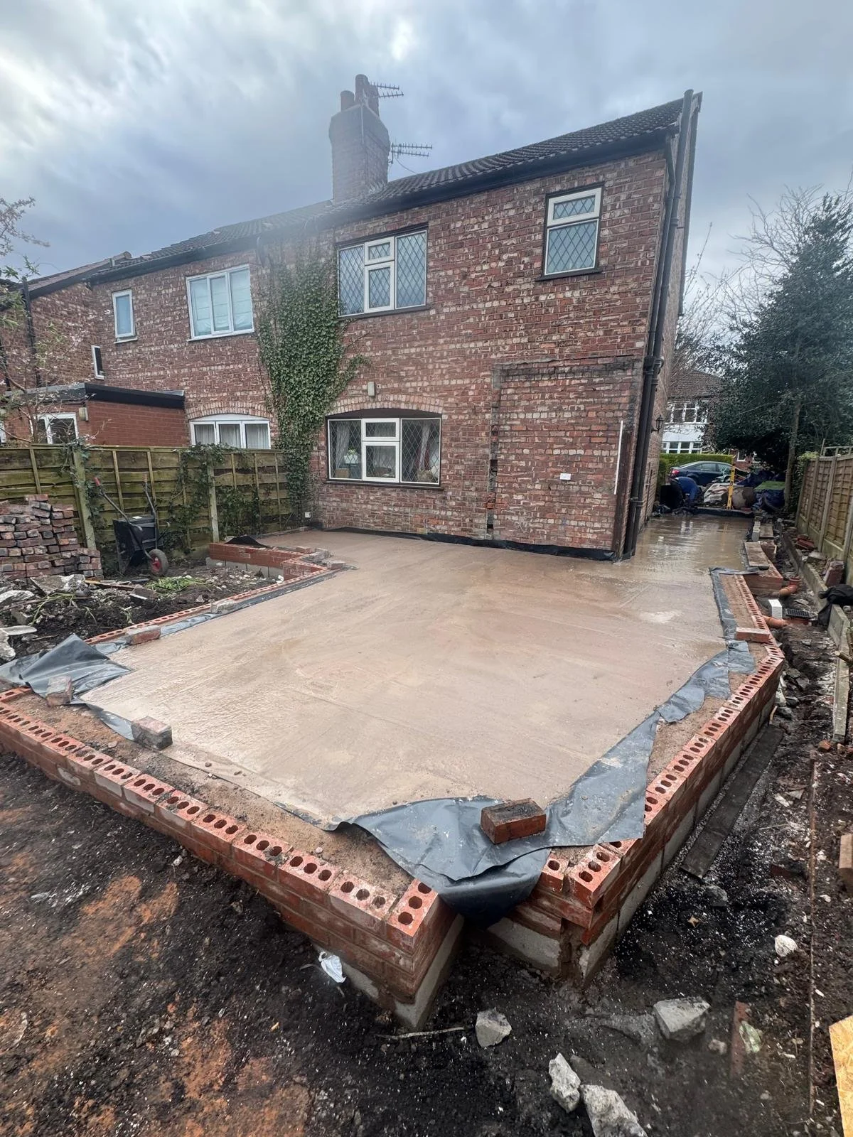 Front yard under construction with leveled concrete slab, brick border, wooden fence on both sides, and a brick house in the background.