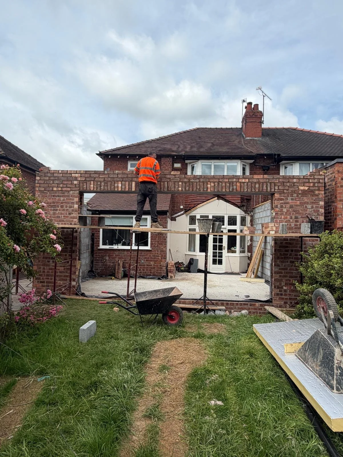 Construction worker in an orange safety vest stands on a temporary wooden platform working on a brick wall extension at a backyard renovation site. The scene shows brick walls, construction tools, and a partly built backyard patio area with a view of