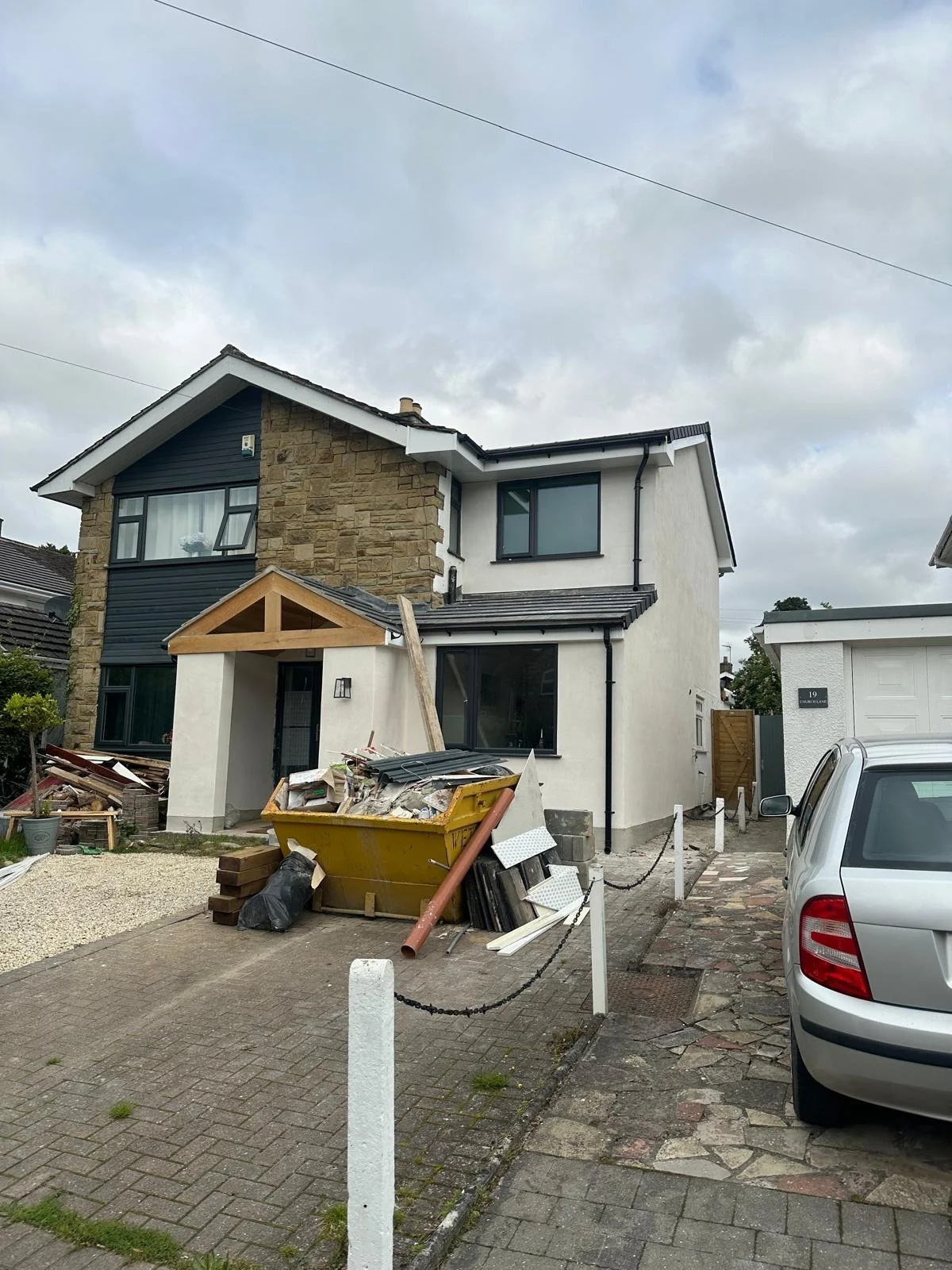 A house under renovation with construction materials and debris in the front yard, a yellow skip filled with construction waste, a parked silver car, and a cloudy sky overhead.