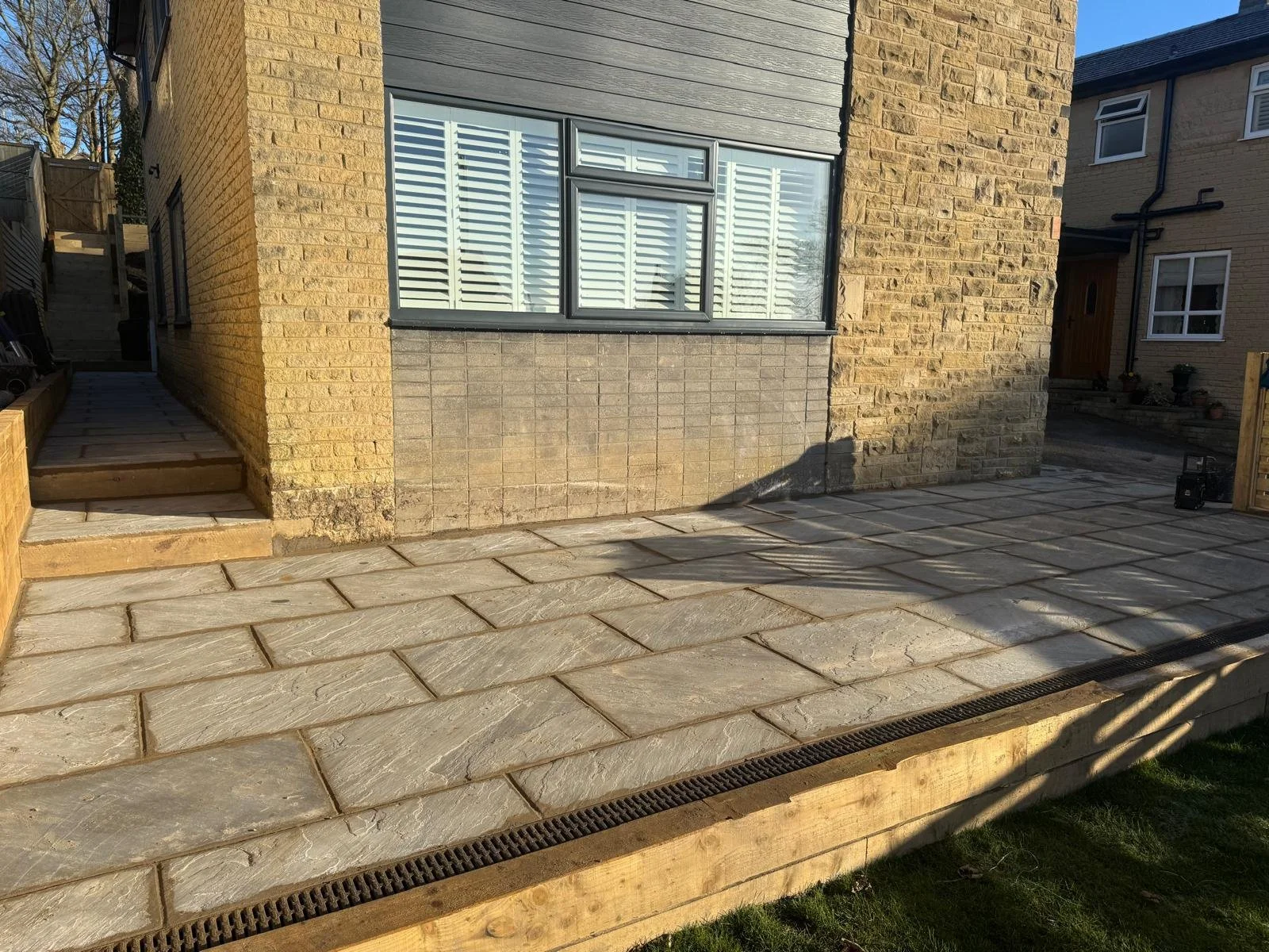 Photo of a newly constructed outdoor patio with large stone tiles, a drainage grate at the edge, and a house with brick and siding exterior in the background. The patio is adjacent to stairs leading up to a door on the house.