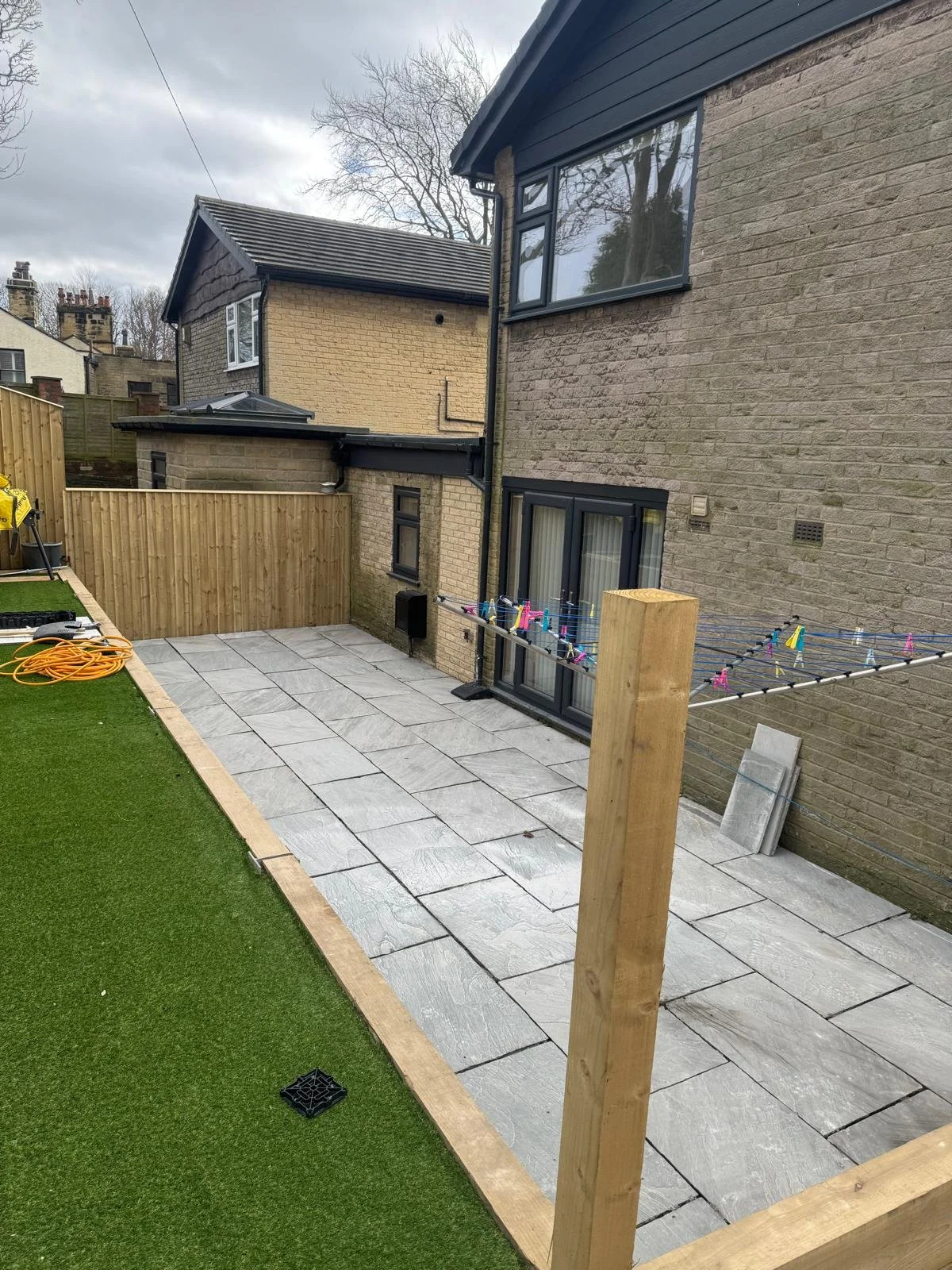 Backyard patio under construction with stone tiles, a wooden fence, and a clothesline with colorful clothespins, next to a lawn.