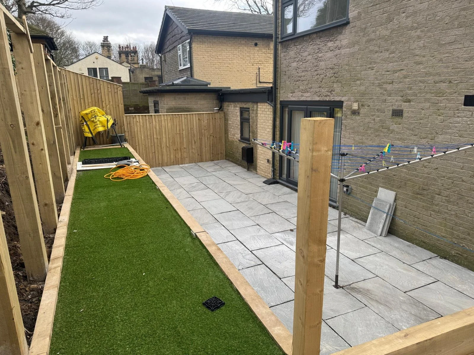 A backyard area under construction with a wooden fence, a section of artificial grass, and paving stones. There is a rotary clothesline with colorful clothespins, a yellow bag, orange extension cord, and construction tools and materials.