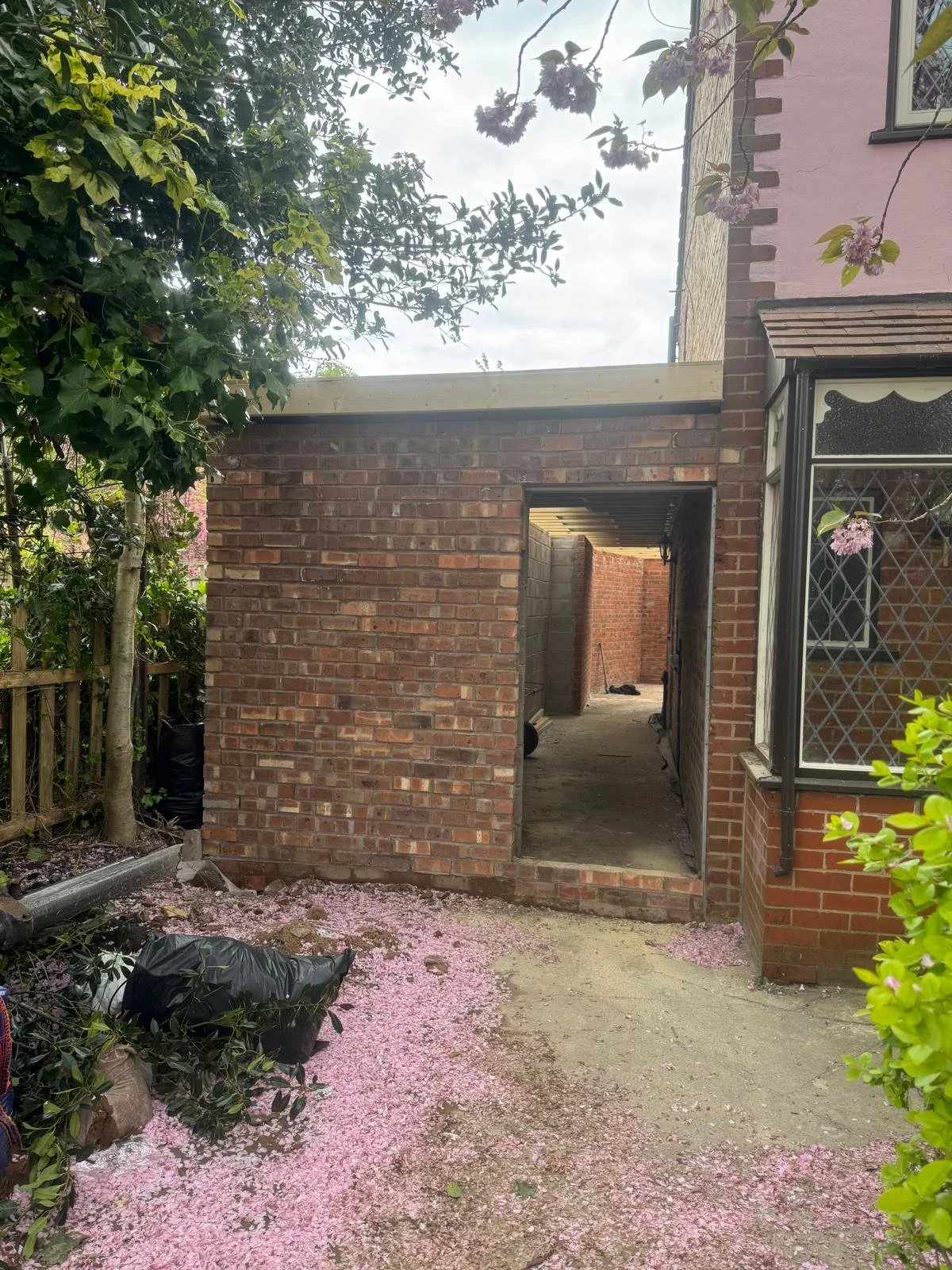 A brick wall with an open doorway under construction, surrounded by greenery and pink flowers on the ground and on a nearby tree.
