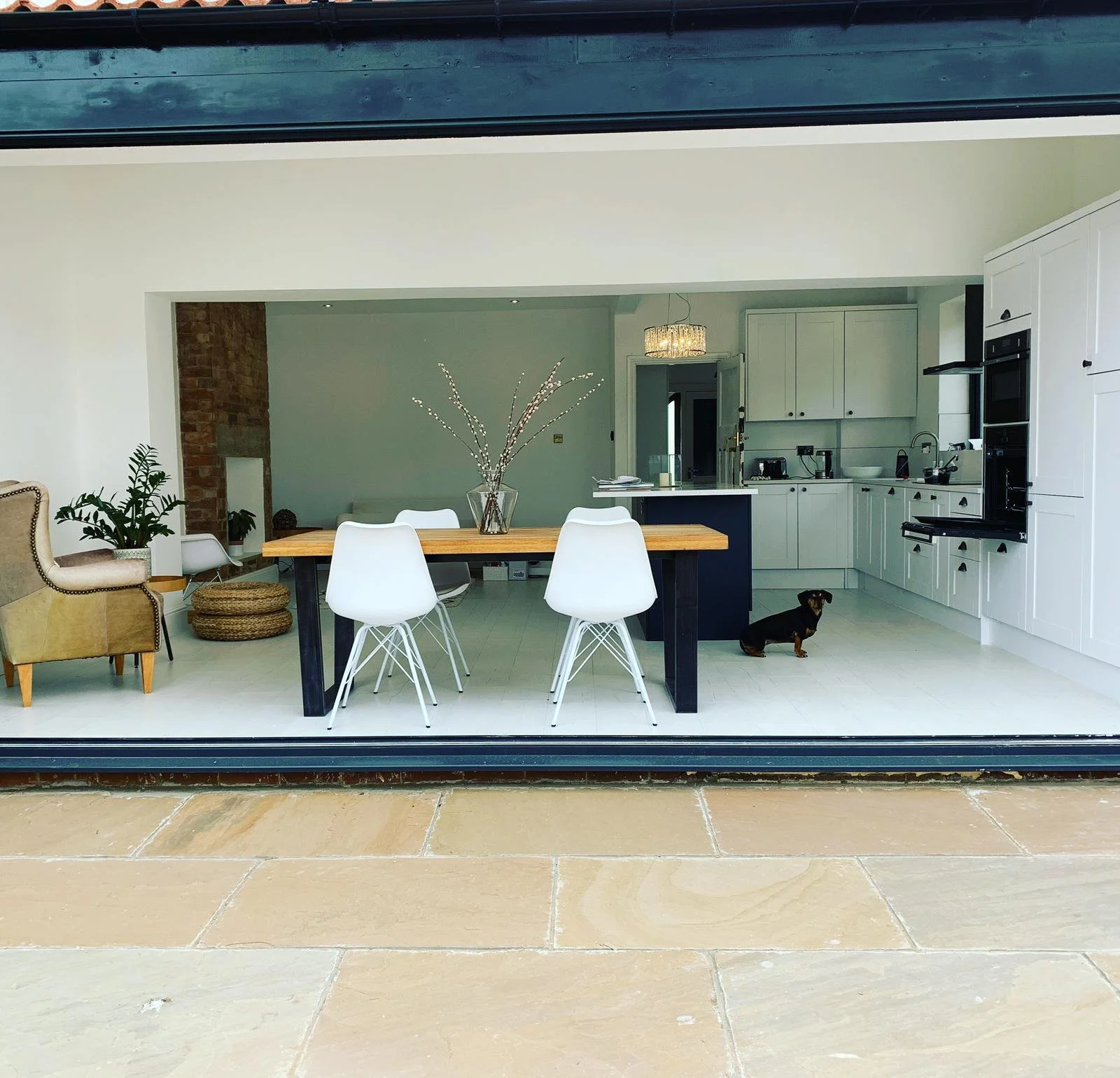 View of a modern kitchen and dining area through a window. The kitchen has white cabinets, a black oven, and a small countertop with a sink. The dining area features a wooden table with four white chairs and a glass vase with branches on top. A small