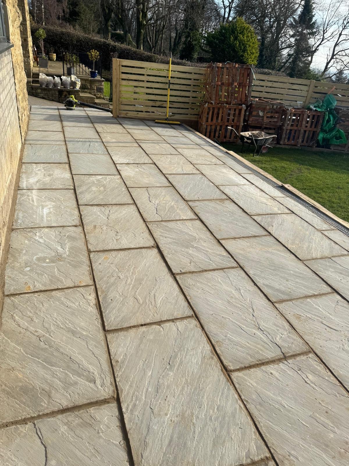 Newly installed rectangular stone patio with textured, cream-colored tiles and dark grout lines, adjacent to a house wall, in a backyard with a wooden fence, grass, and garden tools.
