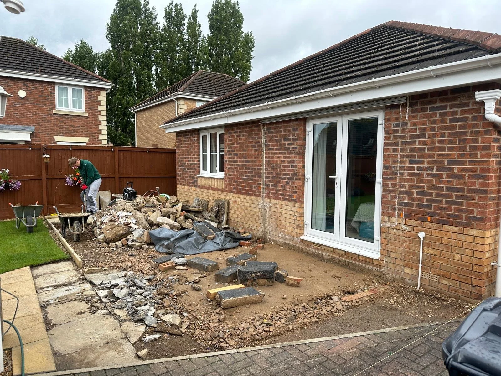 A person working on a backyard renovation, tearing out old paving and digging soil, with construction tools and rubble on the ground, next to a brick house with a sliding glass door and window. There is a wooden fence and neighboring houses in the ba