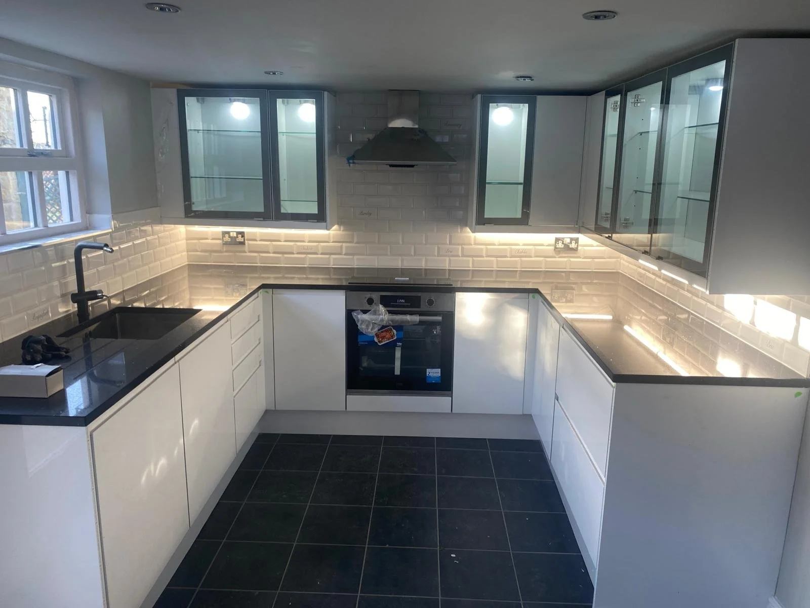 Modern kitchen with white cabinets, black countertops, a black sink, a built-in oven, and glass-front upper cabinets. Under-cabinet lighting and a gray tiled backsplash are visible. There is a window on the left side and ceiling lights.