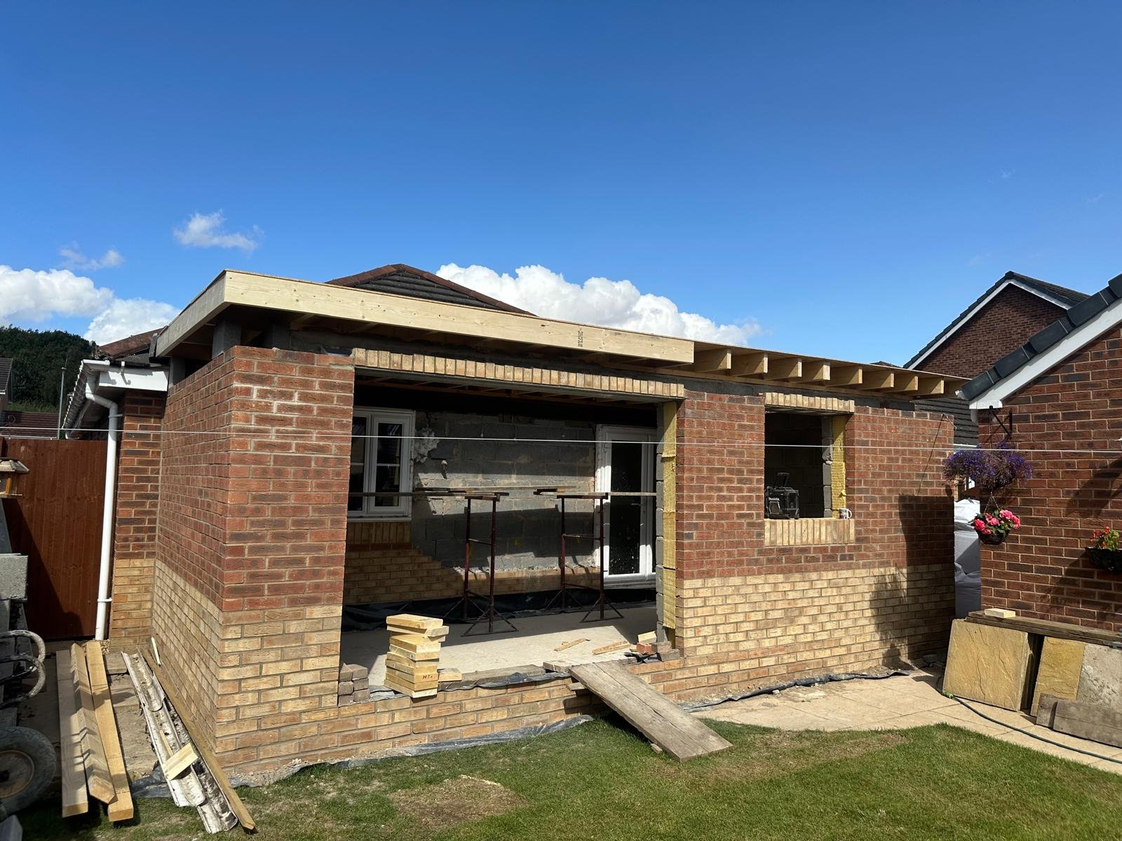 Back of house under construction with brick walls and wooden framing for a new roof extension, construction materials and tools visible.