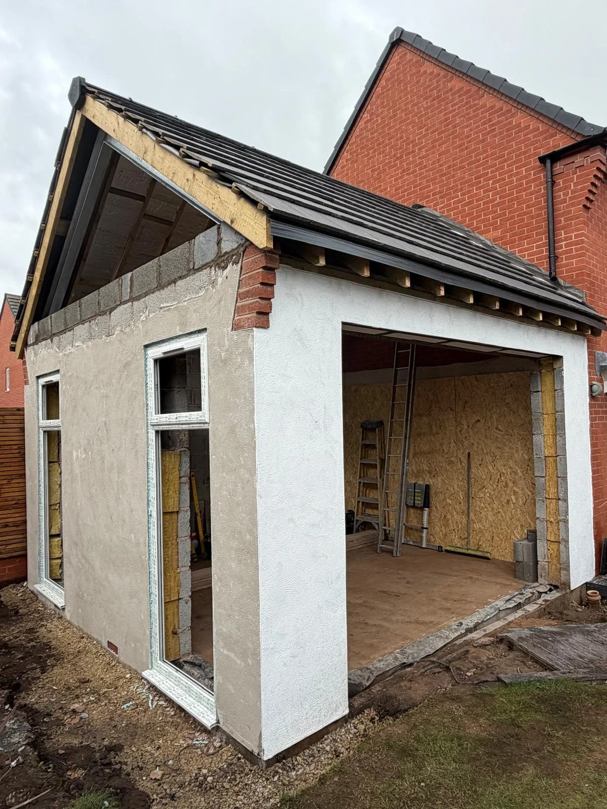 Under construction small house attached to a larger brick house, with an open front, unfinished walls, and construction tools inside.