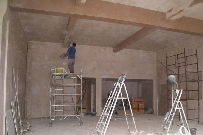 A person in a blue shirt and khaki shorts is working on scaffolding inside a large, unfinished room with beige walls and ceiling, surrounded by construction tools and equipment.
