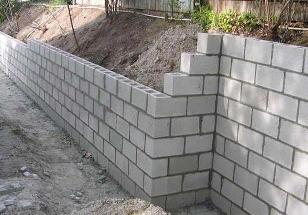 Construction site with a partially built cinder block wall on a dirt surface, with soil and some plants visible behind the wall.