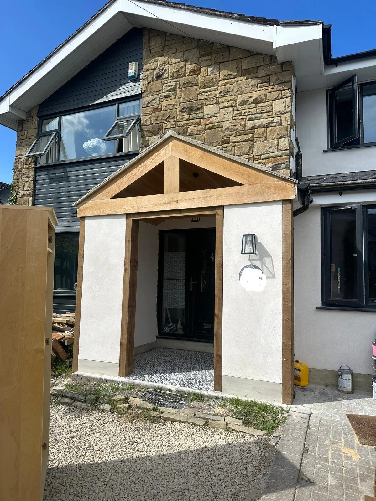 Entrance of a house under construction with a wooden porch frame, black door, outdoor wall sconce, and gravel pathway, with part of the home featuring stone and siding exterior.