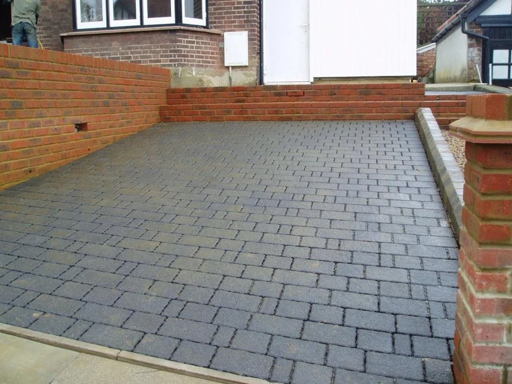A newly paved small outdoor patio with gray bricks, enclosed by red brick walls and located next to a house with a bay window. There is a white door and some brick steps leading up to a different area.