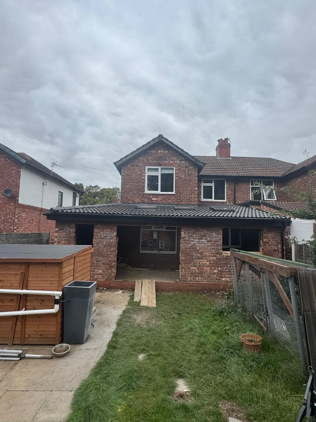 Backyard view of a brick house under construction with an unfinished extension, tools, and construction materials in the yard on a cloudy day.