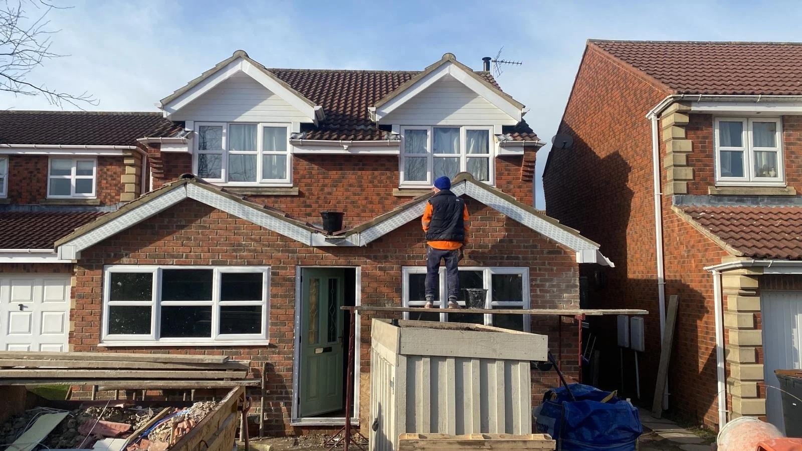 A person working on the roof of a two-story brick house, which is under construction or renovation, with other similar houses nearby.