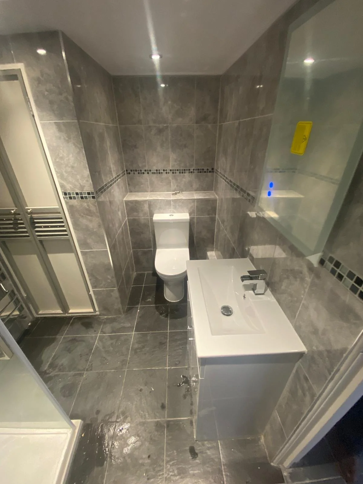 A modern bathroom with gray tiled walls and floor, a toilet, a small sink with a faucet, a mirror, and a shower enclosure, all in a compact space.