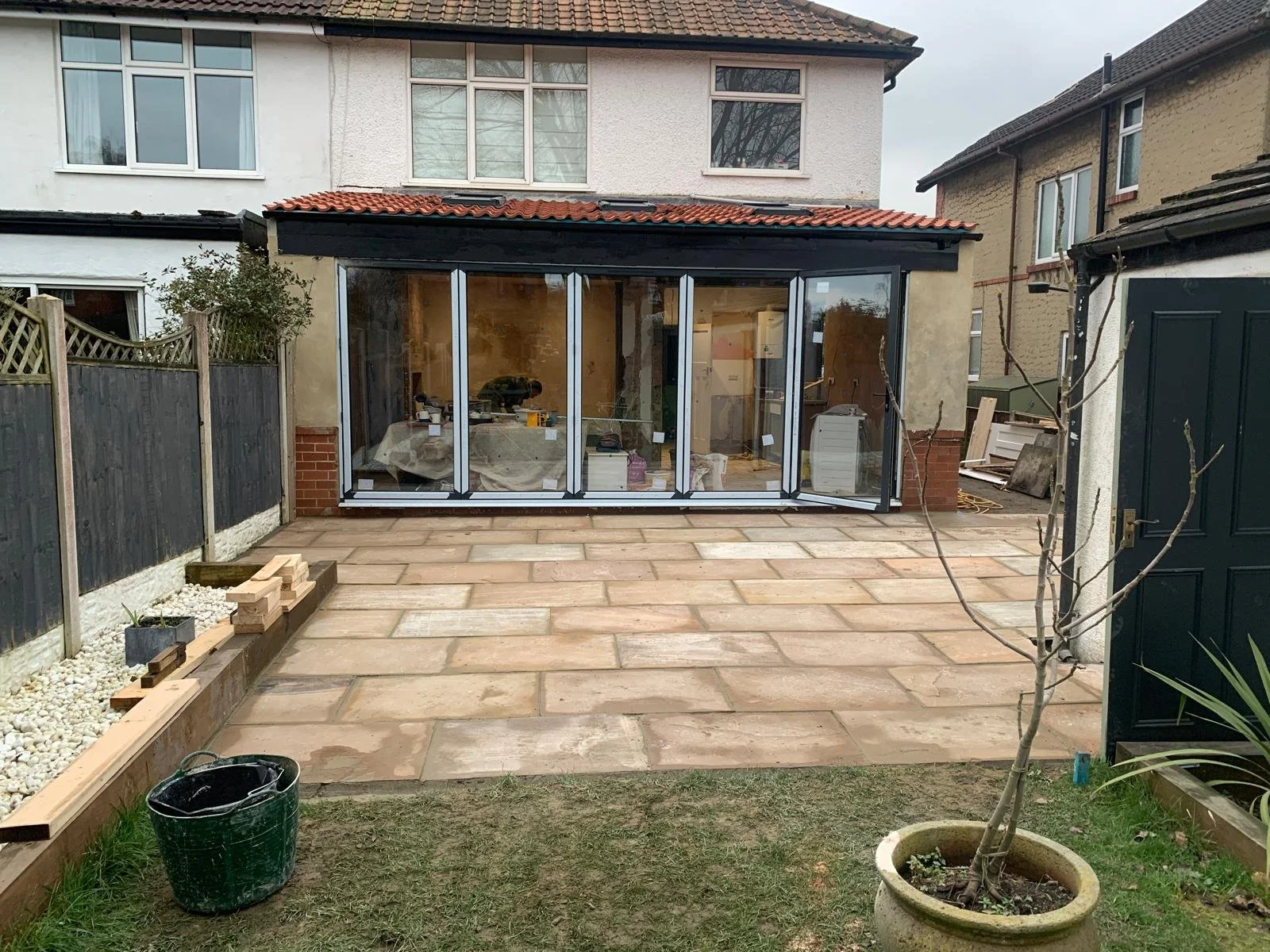 Backyard with a paved patio, small garden bed, and a large glassed-in extension being built onto the house, with tools and construction materials inside.