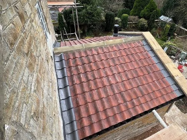 View of a sloped roof with red and gray tiles next to a stone wall, surrounded by trees and garden.