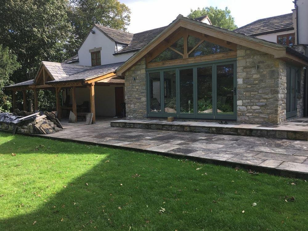 A house with a stone exterior and a large glass sliding door, surrounded by a stone patio and green lawn, with trees in the background.