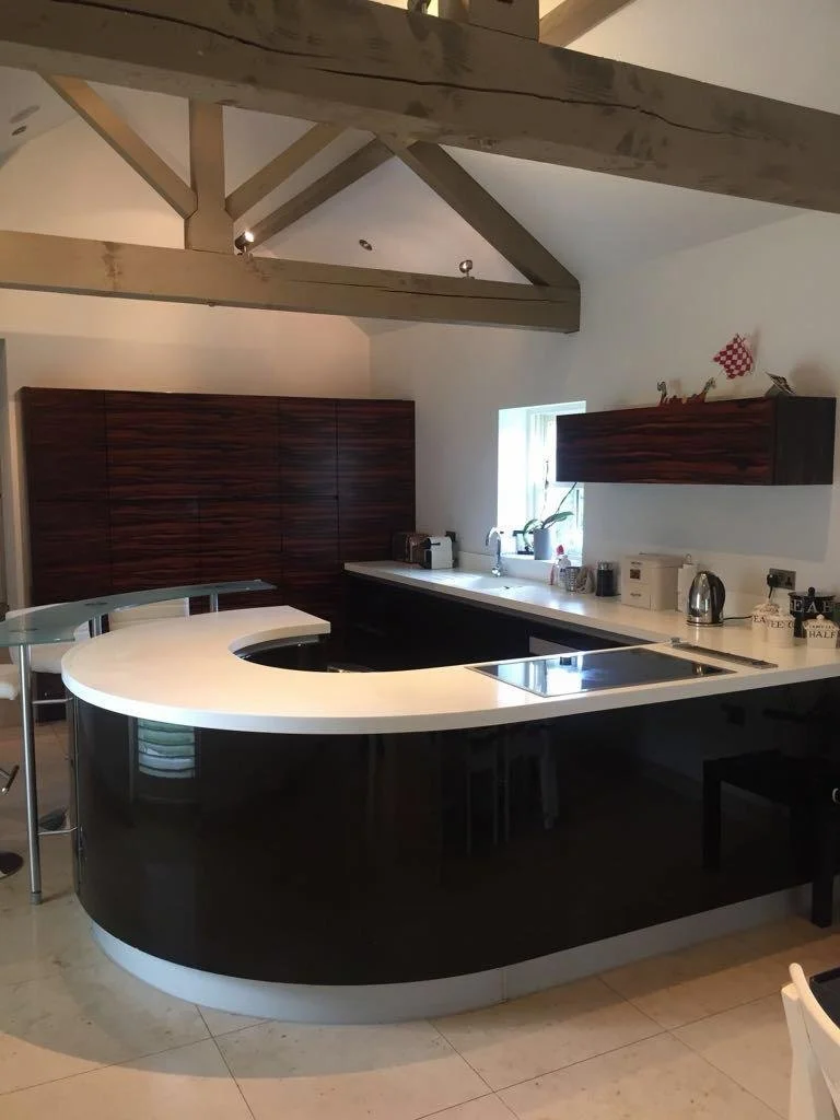 Modern kitchen with curved black island and white countertop, dark wood cabinets, small window, and wooden ceiling beams.