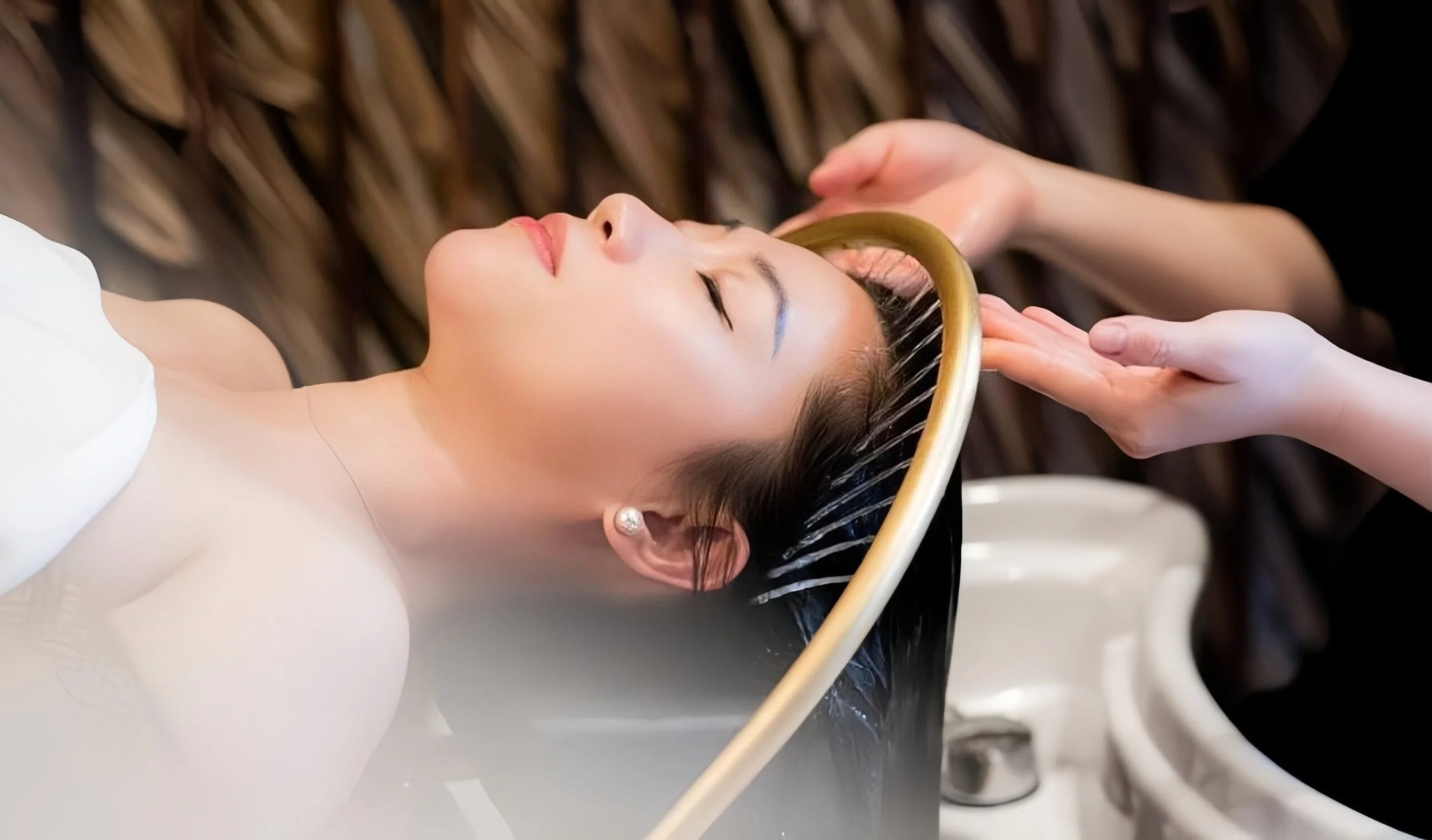 A woman with closed eyes getting her hair washed at a salon, lying on a white sink while a stylist rinses her hair with a yellow hose.