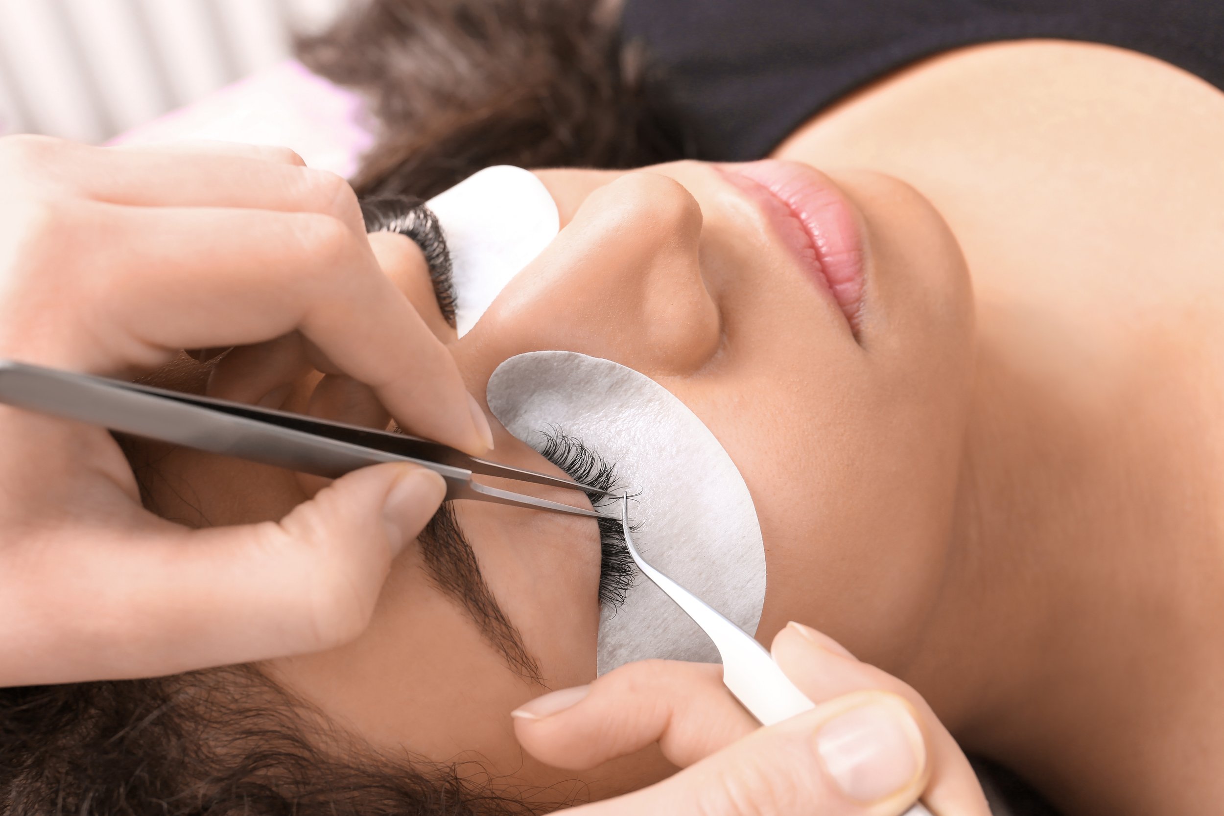 A woman lying down with her eyes closed receives eyelash extensions or makeup application from a professional. The professional is using tweezers to carefully work on her eyelashes while holding a white pad or paper under her eye for protection. The woman has light skin and dark hair, and she appears relaxed.