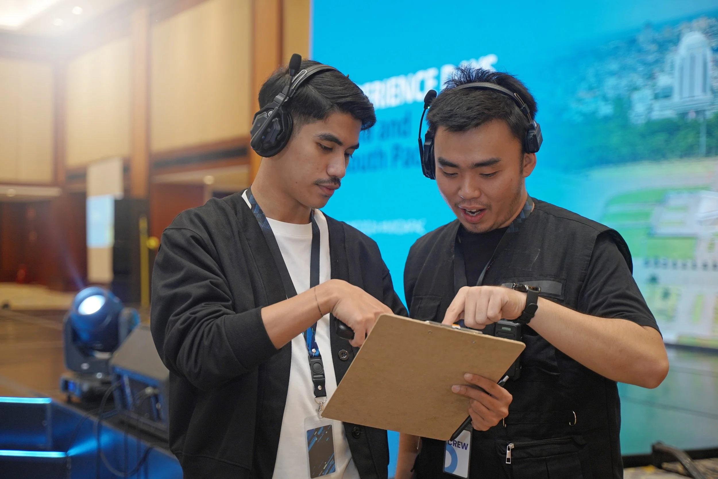 Two young men wearing black jackets and headsets are looking at a tablet together at a conference or event. One is holding the tablet, and the other is pointing at it.