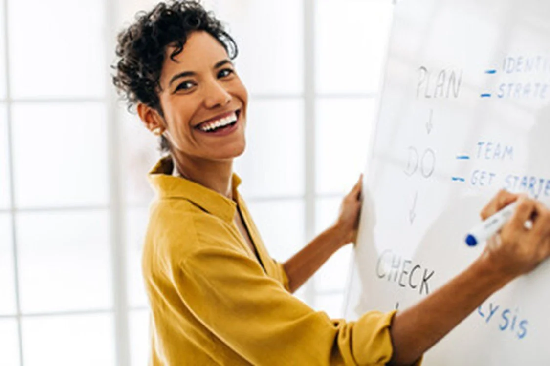 A woman in a yellow shirt smiling as she writes on a whiteboard with notes and diagrams.