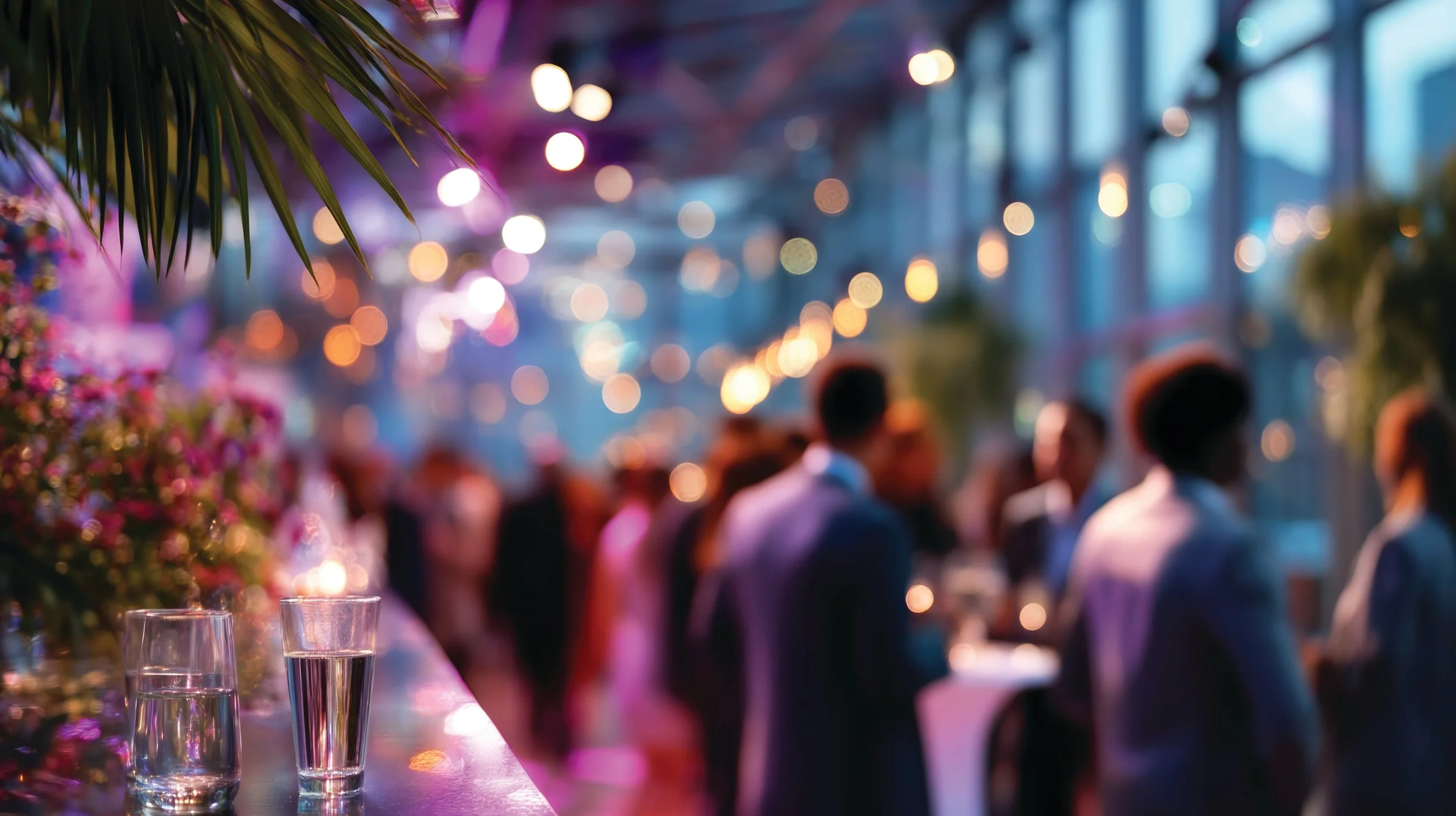 Blurred scene of a crowded social event or party with string lights hanging overhead, pink flowers on the left, and glasses of water in the foreground.