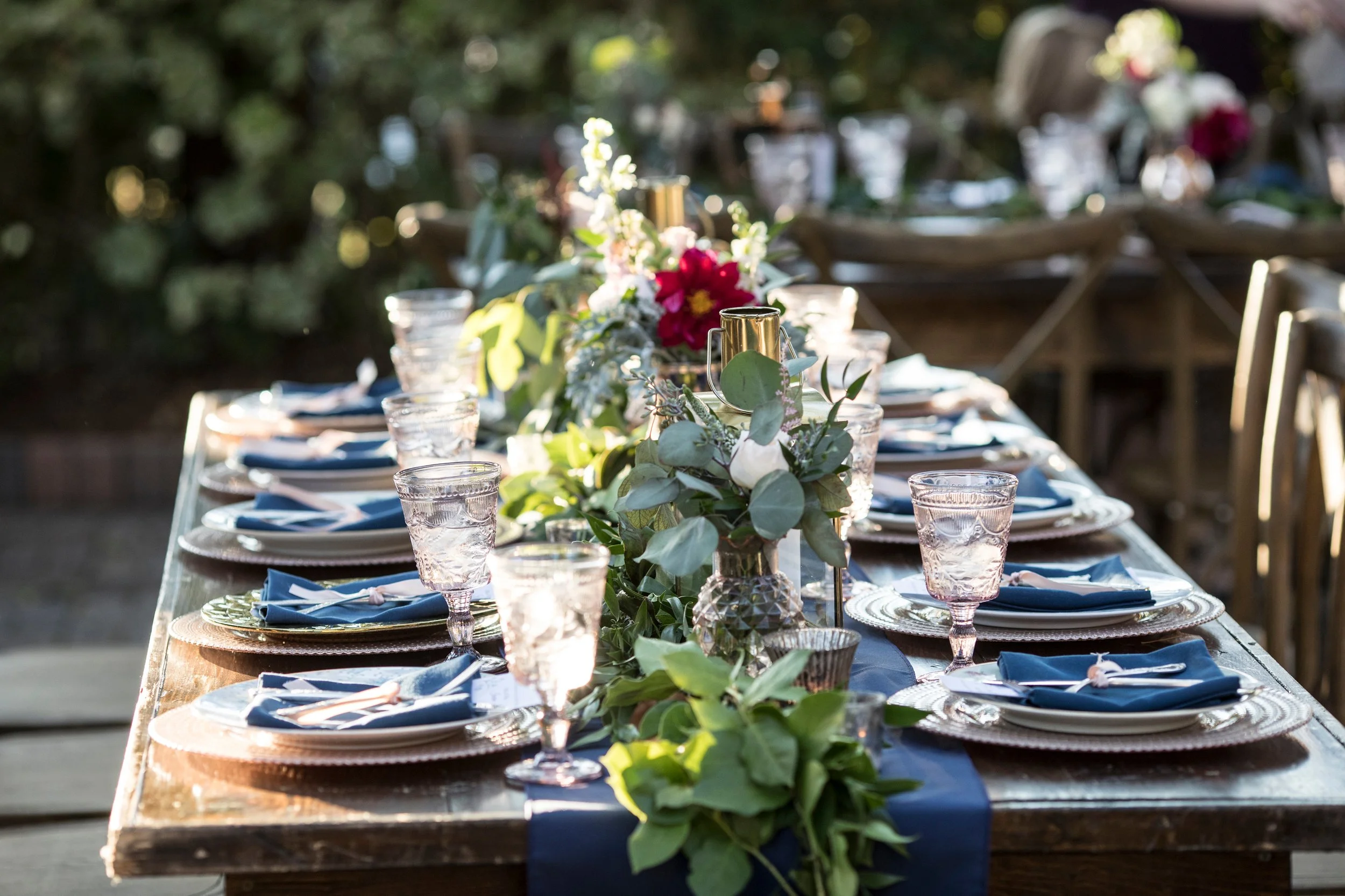 Outdoor banquet table decorated with a lush green garland, pink and white flowers, gold candle holders, and pink glassware, set for a celebration.