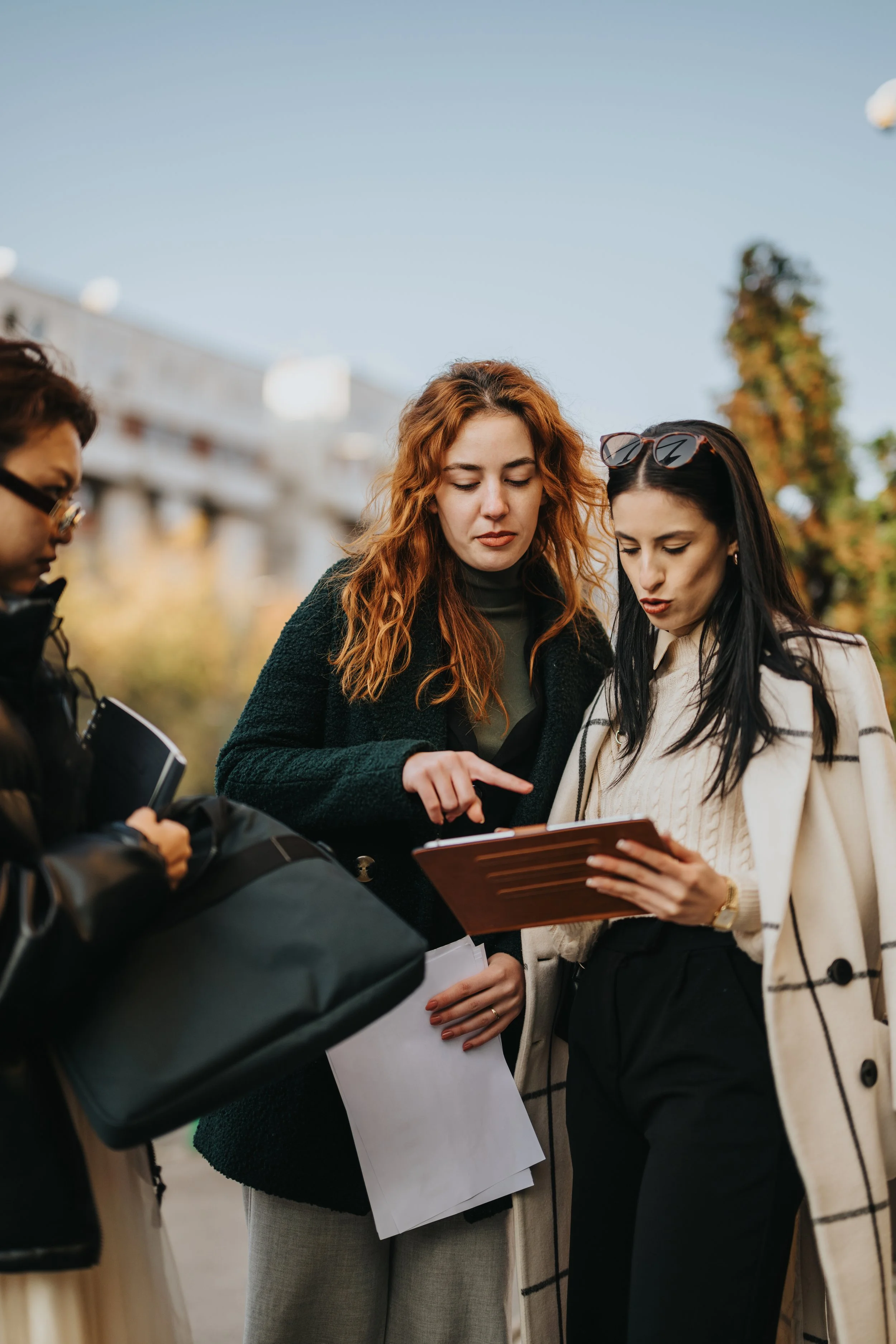 Group of people outdoors, looking at a document or device, with trees and buildings in background, possibly in a city park or downtown area.
