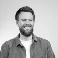 Et sort-hvid portrætfoto af en mand med skæg, iført en skjorte og en T-shirt, der smiler.