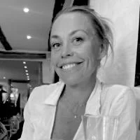 En smilende kvinde med lyst hår, iført hvidt tøj, sidder ved et bord i en restaurant eller café.