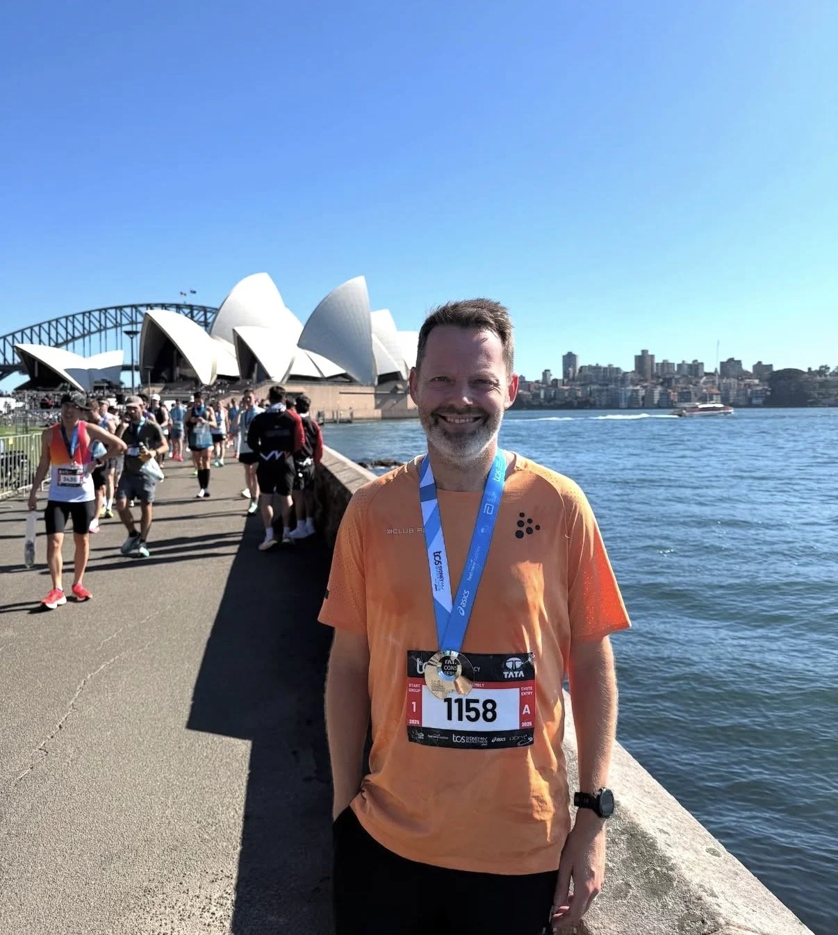 En mand smiler foran operahuset i Sydney med en medalje om halsen efter et løb. Der er andre løbere i baggrunden, og byen Sydney kan ses i baggrunden.