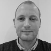 Sort/hvid portrætfoto af en mand med kort hår og let smil, iført en skjorte og sweater.