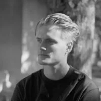 Sort-hvid foto af en ung mand med blond hår, der står udenfor med træer i baggrunden.