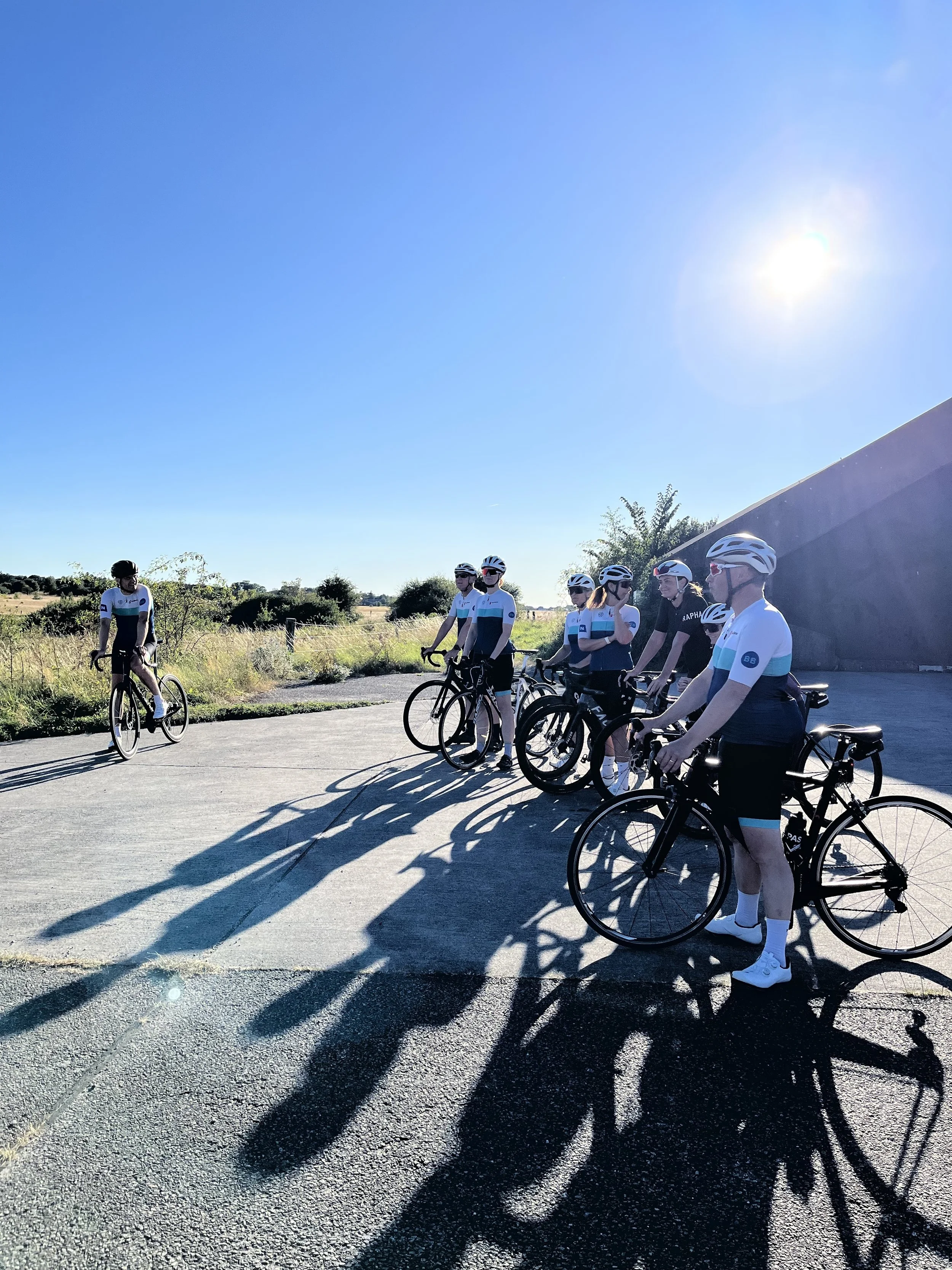 En gruppe cykelryttere står sammen udendørs i sollys, nogle vinker, og de har cykler. Solen skinner klart over dem.