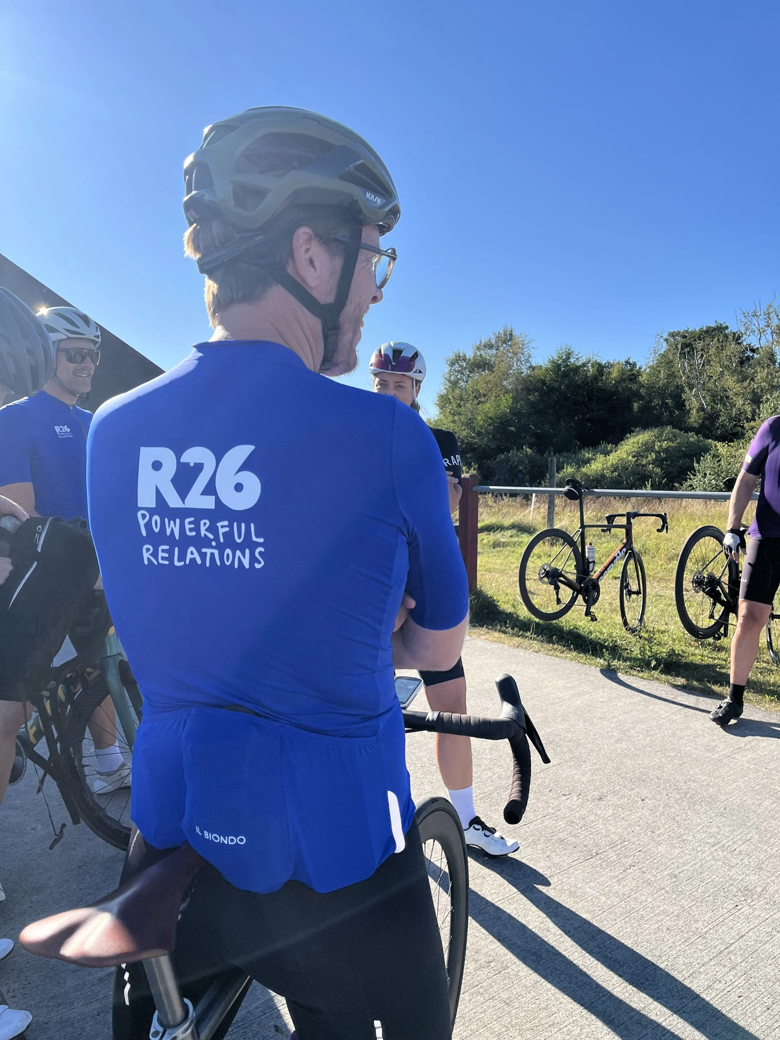 Gruppe cyklister i cykeludstyr, inklusive hjelme, mødes ude i det grønne på en solrig dag