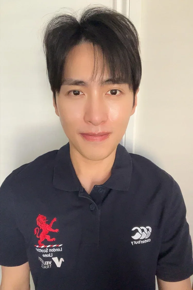 A young man with dark hair, wearing a black polo shirt with logos, smiling at the camera.