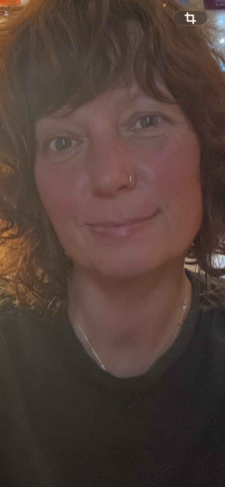 Close-up of a woman with curly auburn hair, a nose piercing, and a gentle smile, wearing a black top and a delicate necklace.