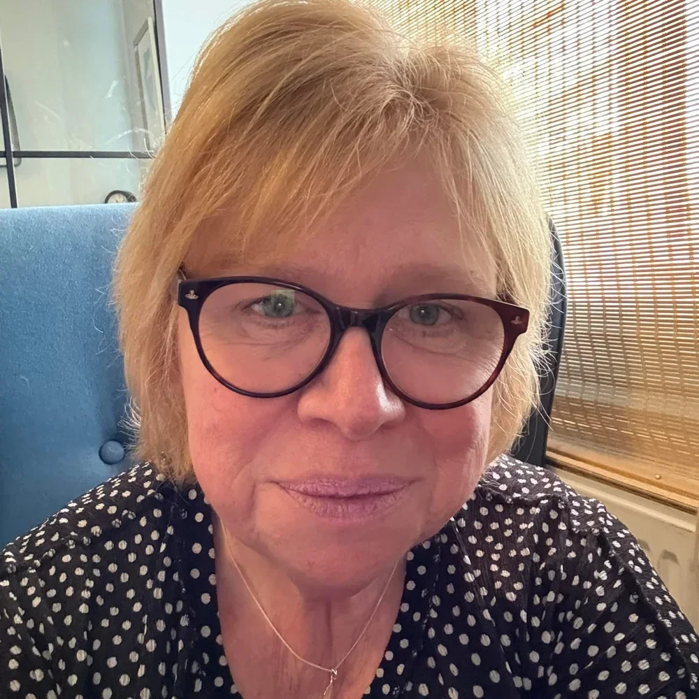 Close-up of a woman with short blonde hair, wearing glasses and a black blouse with white polka dots, sitting in a room with wooden blinds.