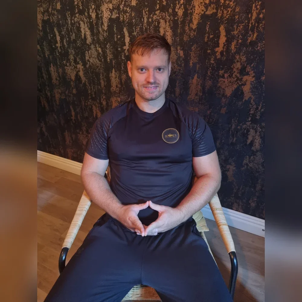 A young man with short brown hair and a beard sitting on a woven chair in front of a textured dark wall. He is wearing a black athletic shirt with a circular logo on the chest and black pants. The man is smiling and has his hands clasped together in front of his stomach.