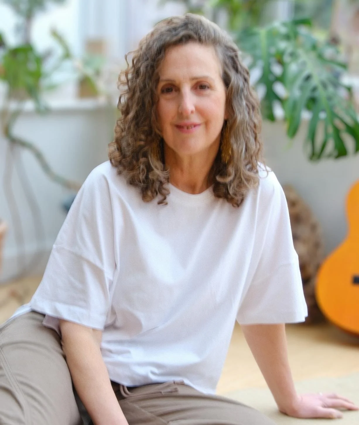 A woman with curly brown hair, wearing a white t-shirt and beige pants, sitting on the floor in a brightly lit room with green plants in the background.