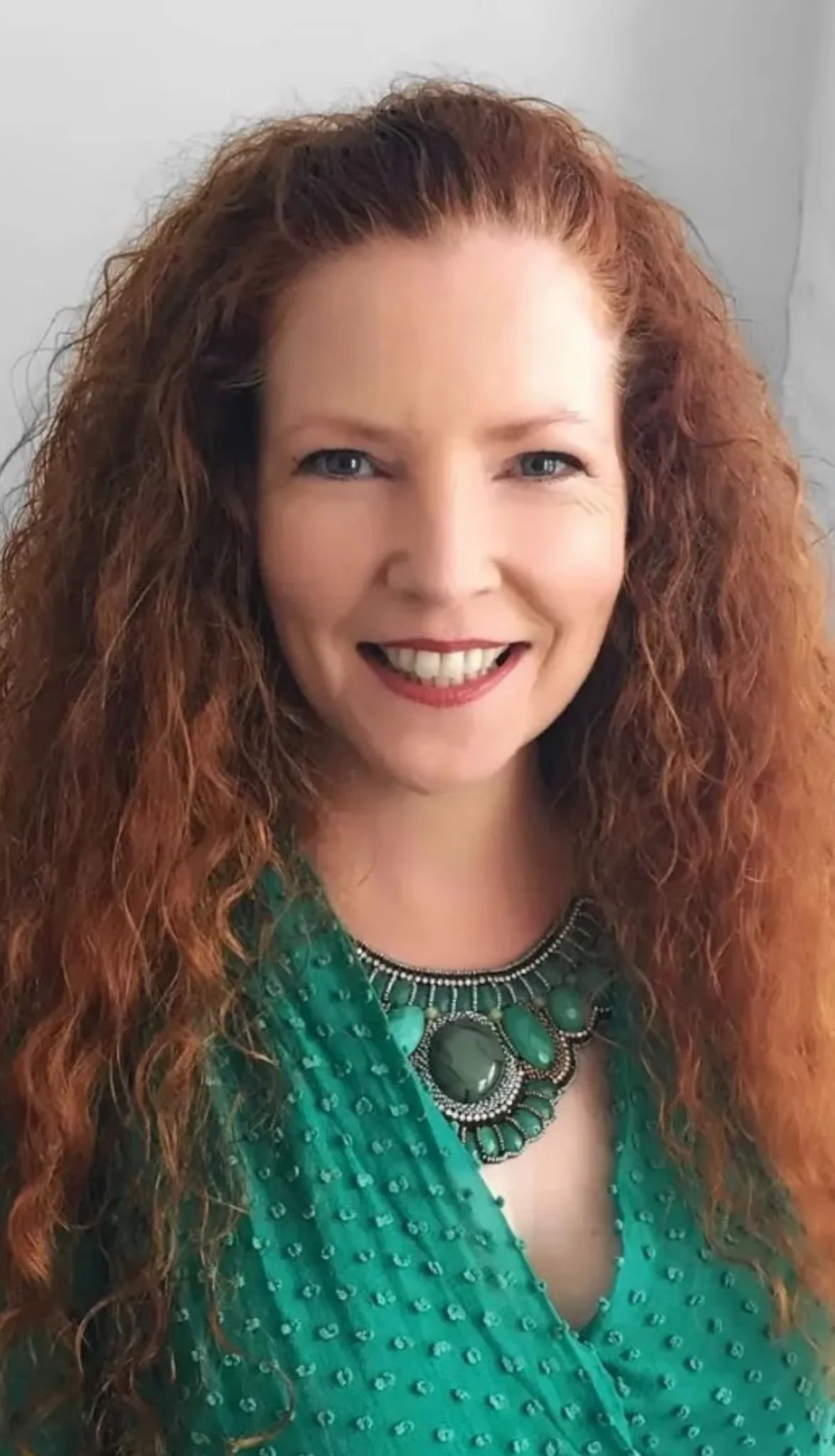 Close-up of a smiling woman with long curly red hair, blue eyes, wearing a green patterned dress and a large statement necklace.