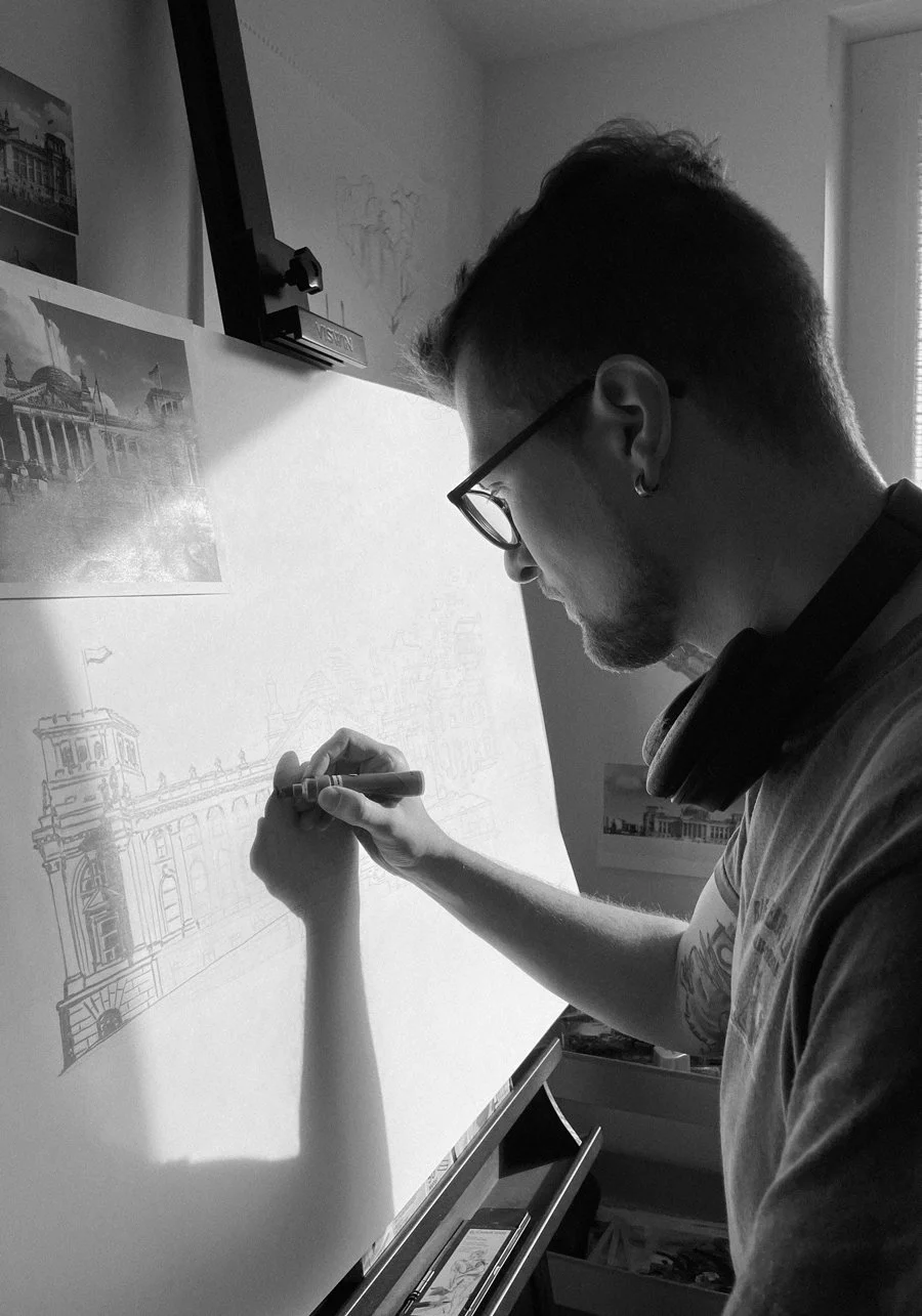 A black and white photo of youthful artist with glasses is drawing an architectural building, with a reference photo pinned to the wall behind him.