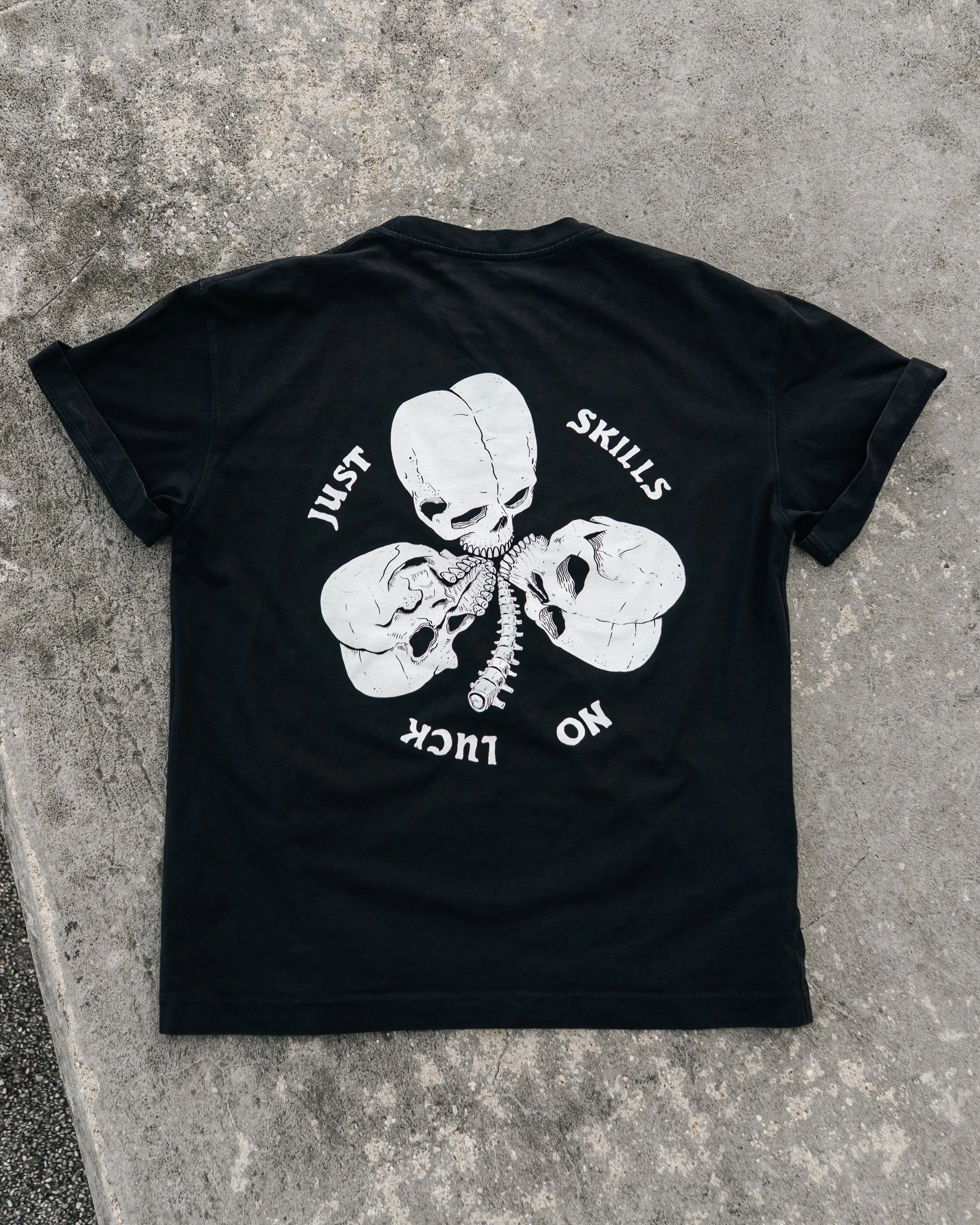 A black T-shirt lying flat on a concrete surface. The T-shirt has a white graphic of a three skulls on it and with the words "Just Skills, No Luck".